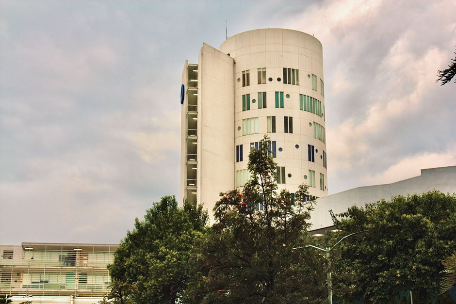 Contemporary white cylindrical building surrounded by trees under a cloudy sky in Ciudad López Mateos, Mexico.